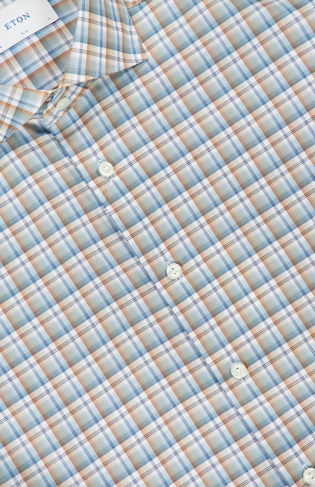 A close-up of the Check Twill Slim Dress Shirt by ETON, showcasing its slim fit design and plaid pattern in light blue, tan, white, and orange. Crafted from wrinkle-resistant organic cotton, this dress shirt features a light blue checkered print with four white buttons visible along with part of the collar and the signature ETON label.