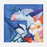 The ETON Floral Oxford Silk Pocket Square features vibrant blue, purple, white, orange, and yellow floral tones on dark blue silk. The word ETON is near the top left, and one corner is folded to reveal its reverse side.