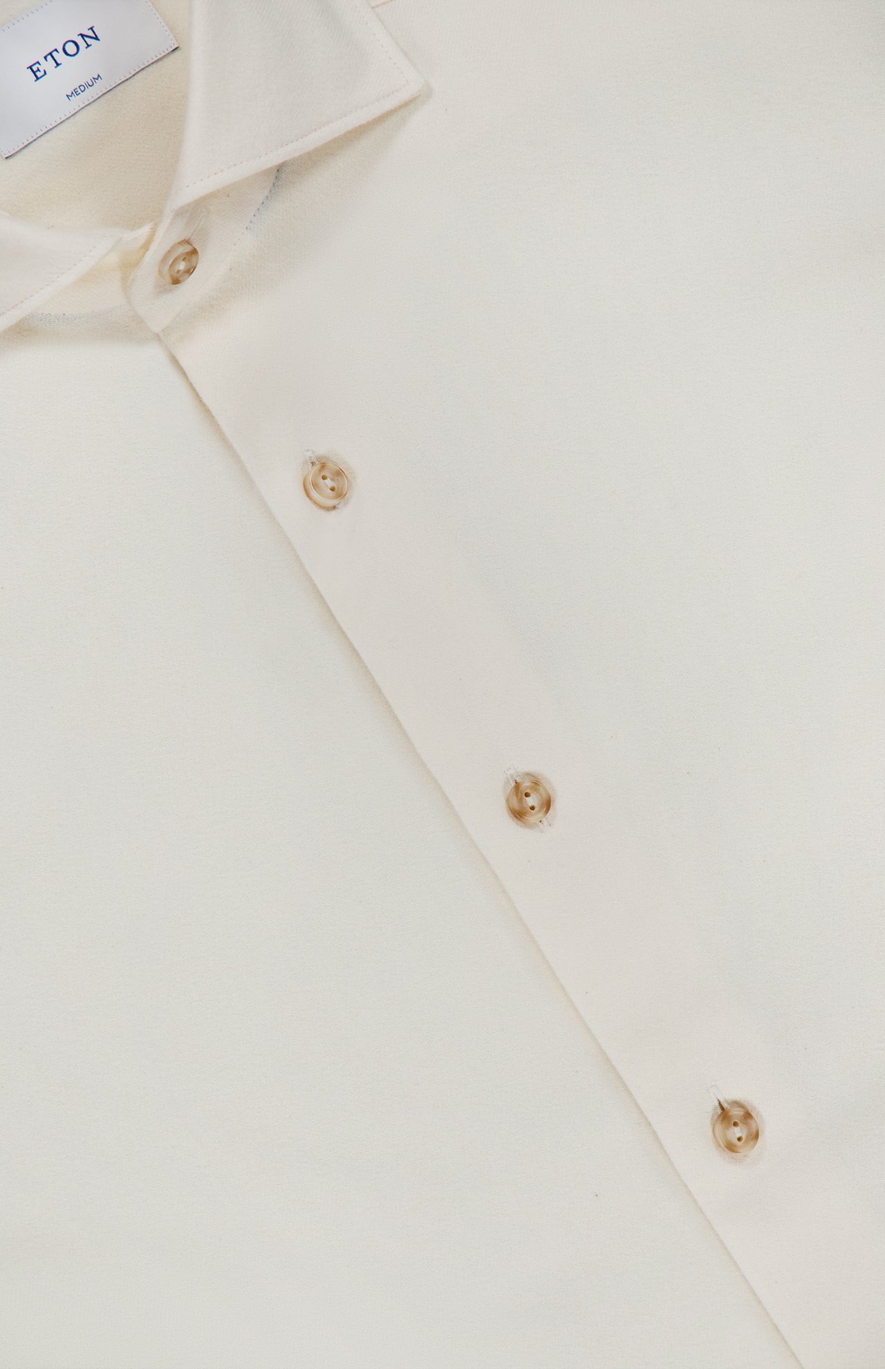 An off-white Solid Flannel Shirt by ETON, featuring beige buttons and a collar tag that displays “ETON” and “MEDIUM,” is shown neatly folded on a flat surface. Designed for casual comfort, this cotton shirt exudes effortless style.