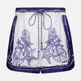 The Coast Scarf Shorts by FARM RIO are white coastal vacation shorts with a navy waistband, blue floral and leaf embroidery on the front, borders, and a braided drawstring tie at the waist.