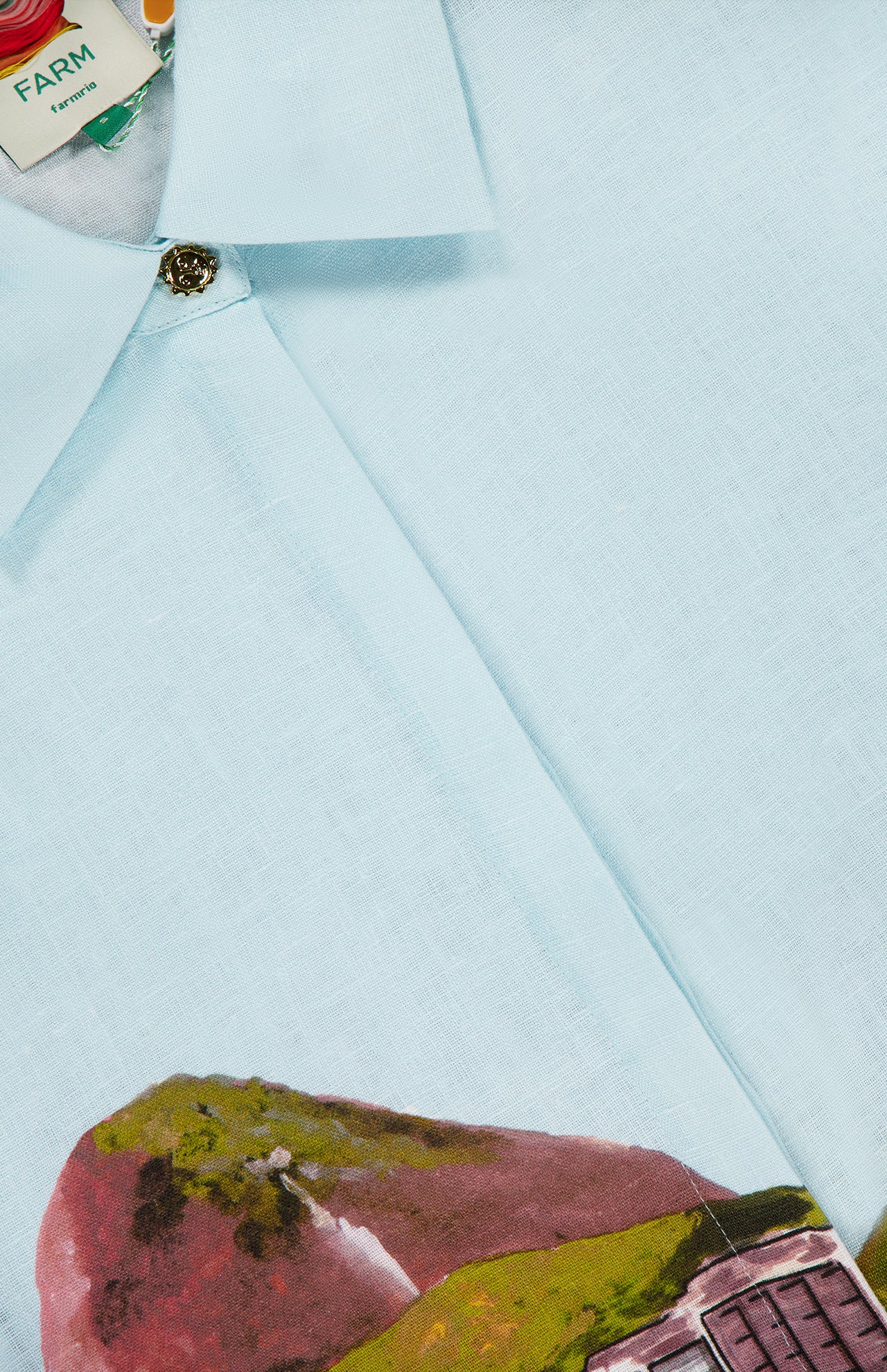 Close-up of the light blue Copacabana Beach Long Sleeve Kimono by FARM RIO, featuring a button and vibrant landscape print at the hem. The collar tag displays FARM, alongside mountain and building imagery.