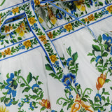 A close-up of the FARM RIO Emily Floral Tapestry Mini Dress, featuring white fabric with vibrant green, blue, yellow, and orange floral and pineapple embroidery and a blue-trimmed waist tie.