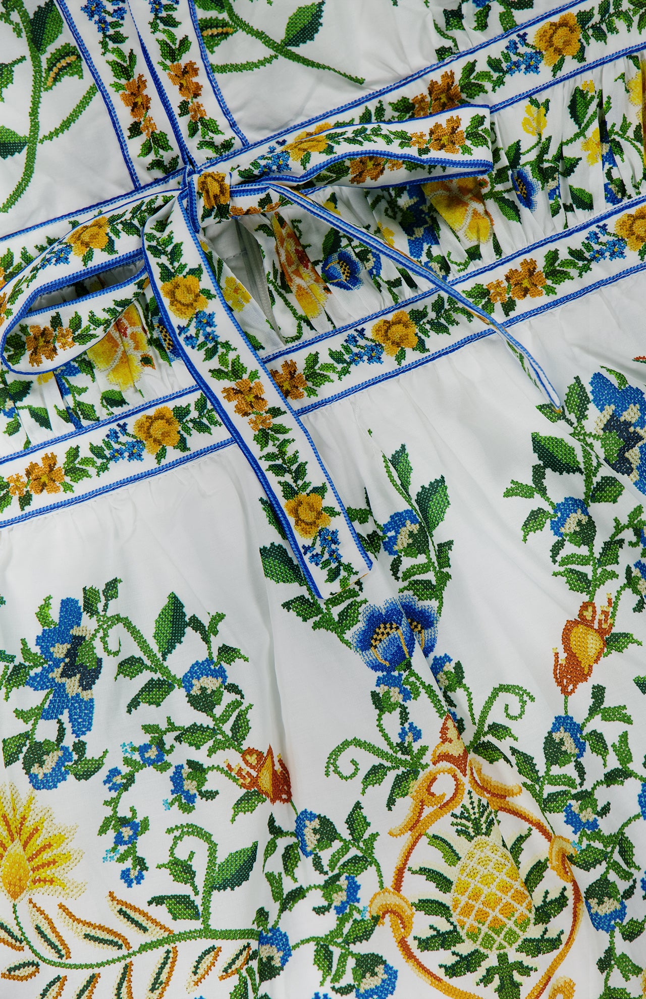 A close-up of the FARM RIO Emily Floral Tapestry Mini Dress, featuring white fabric with vibrant green, blue, yellow, and orange floral and pineapple embroidery and a blue-trimmed waist tie.