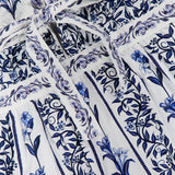 Close-up of the FARM RIO Flowers Arch Mini Dress, showcasing blue floral and leaf motifs over white with vertical stripes, a tied waist belt, decorative borders, and a delicate ruffled hem.