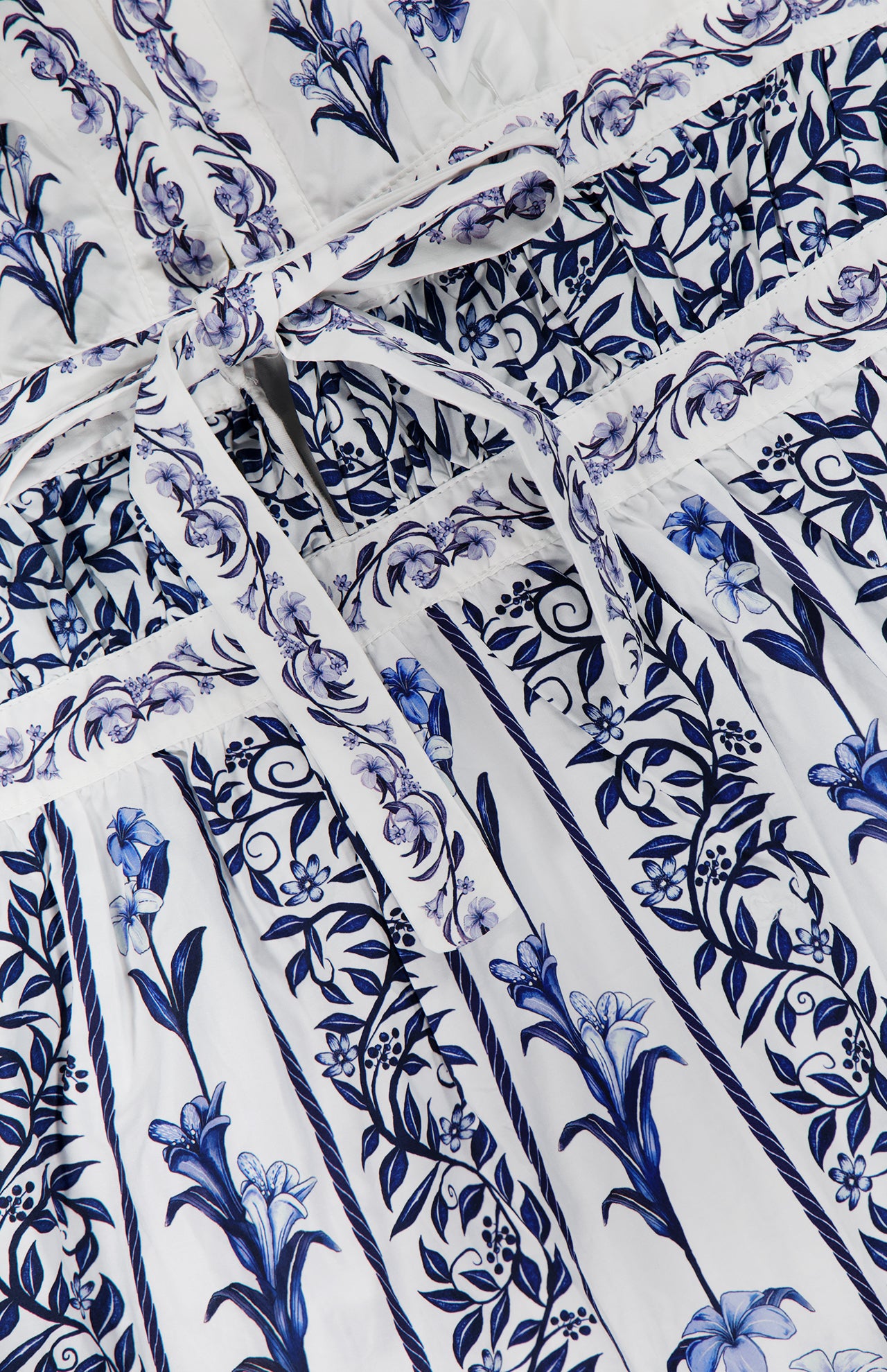 Close-up of the FARM RIO Flowers Arch Mini Dress, showcasing blue floral and leaf motifs over white with vertical stripes, a tied waist belt, decorative borders, and a delicate ruffled hem.