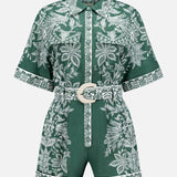 The Miranda Short Sleeve Belted Romper by FARM RIO features green fabric with white floral patterns, a collared neckline, front buttons, and a matching belt with a round buckle. Floral trim accents the hem and button placket.