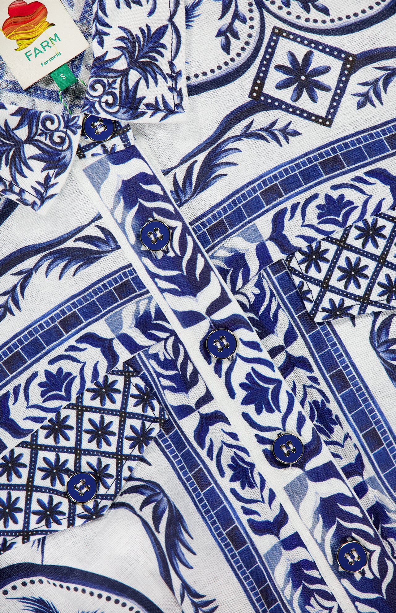 Close-up of the FARM RIO Wonderful City Linen Romper, featuring blue and white nature-inspired patterns and blue buttons down the front, with a fabric tag inside the collar reading FARM RIO.