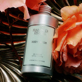 A silver bottle of FLAMINGO ESTATE Jasmine Damask Rose Body Wash sits on a metallic, shell-shaped surface, surrounded by large pink damask roses. The label highlights natural sea salt and a 12 fl oz size.