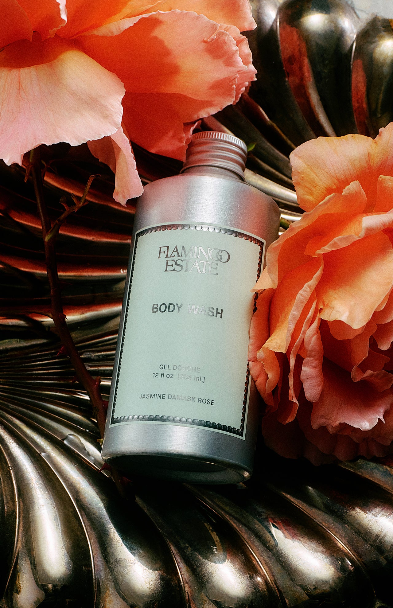 A silver bottle of FLAMINGO ESTATE Jasmine Damask Rose Body Wash sits on a metallic, shell-shaped surface, surrounded by large pink damask roses. The label highlights natural sea salt and a 12 fl oz size.