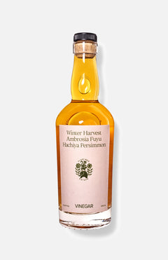 A clear glass bottle of FLAMINGO ESTATE Persimmon Vinegar contains amber liquid and features a tree logo, black text on a cream label reading Winter Harvest Ambrosia Fuyu Hachiya Persimmon, plus a dark cap, set against a white background.