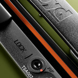 Close-up of The Bold Suitcase's black hard-top lock with combination dial and TSA007 keyhole, showing "FLOYD" embossed on the handle, set against a green background.