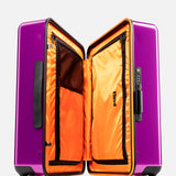 The Cabin Suitcase by FLOYD is a purple hard-top case with black handles, a built-in TSA lock, and red skateboard-style wheels. Open it to reveal a bright orange interior with zippered compartments for organized travel.