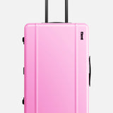 The FLOYD Check-In Suitcase, a hard-top pink case with black handles, a TSA lock, and four red 360° spinner wheels, stands upright against a plain white background.