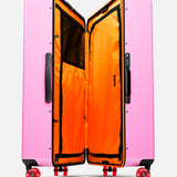 The Trunk Suitcase by FLOYD is a hard-top pink case with a TSA lock, revealed upright with a vibrant orange interior, zippered compartments, black mesh pockets, and red 360° spinner wheels for easy maneuverability.