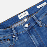 Close-up of premium stretch denim blue jeans displaying the waistband, belt loops, button, front pocket, and FRAME brand label with a tag reading L’Homme Slim Jean in Verdugo. These slim fit jeans by Frame feature classic detailing in a rich dark blue hue.