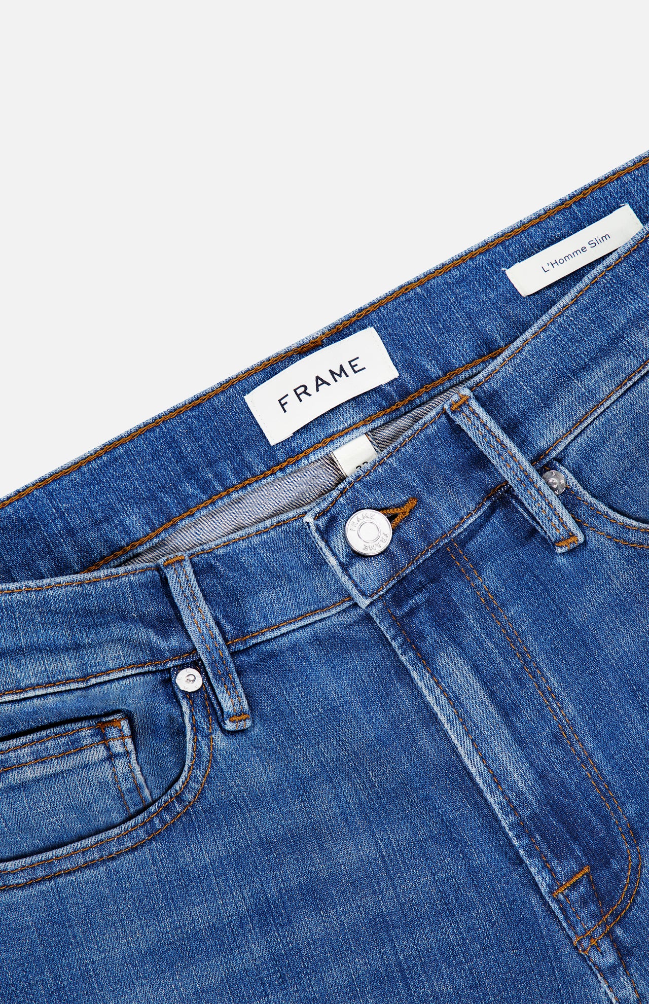 Close-up of premium stretch denim blue jeans displaying the waistband, belt loops, button, front pocket, and FRAME brand label with a tag reading L’Homme Slim Jean in Verdugo. These slim fit jeans by Frame feature classic detailing in a rich dark blue hue.