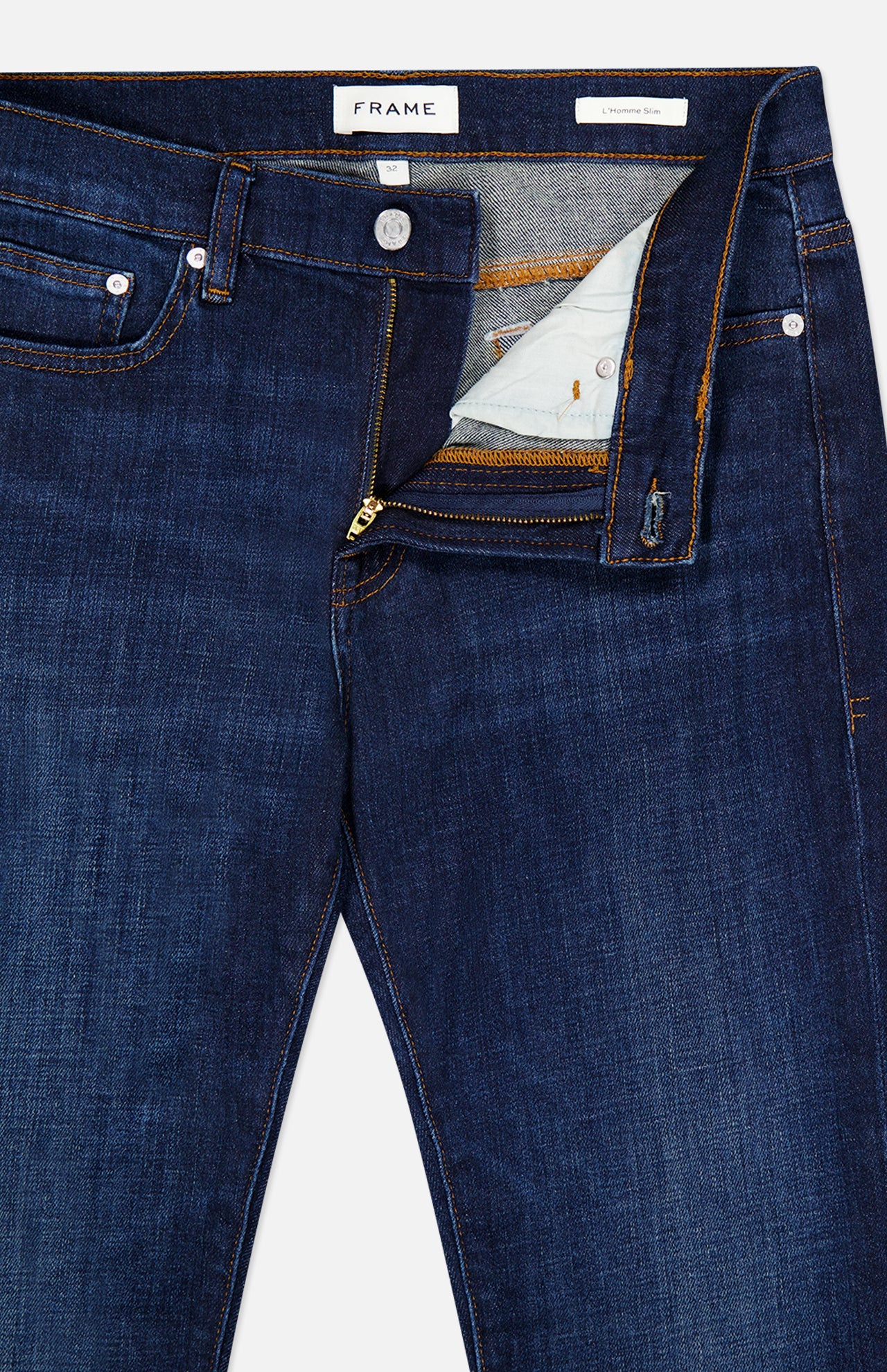 L’Homme Slim Jean in Baltic by Frame, crafted from premium stretch dark blue denim, features a partially open visible zipper fly revealing the inside lining and button. These slim fit jeans are detailed with copper stitching, silver rivets, and a FRAME label on the inside waistband.