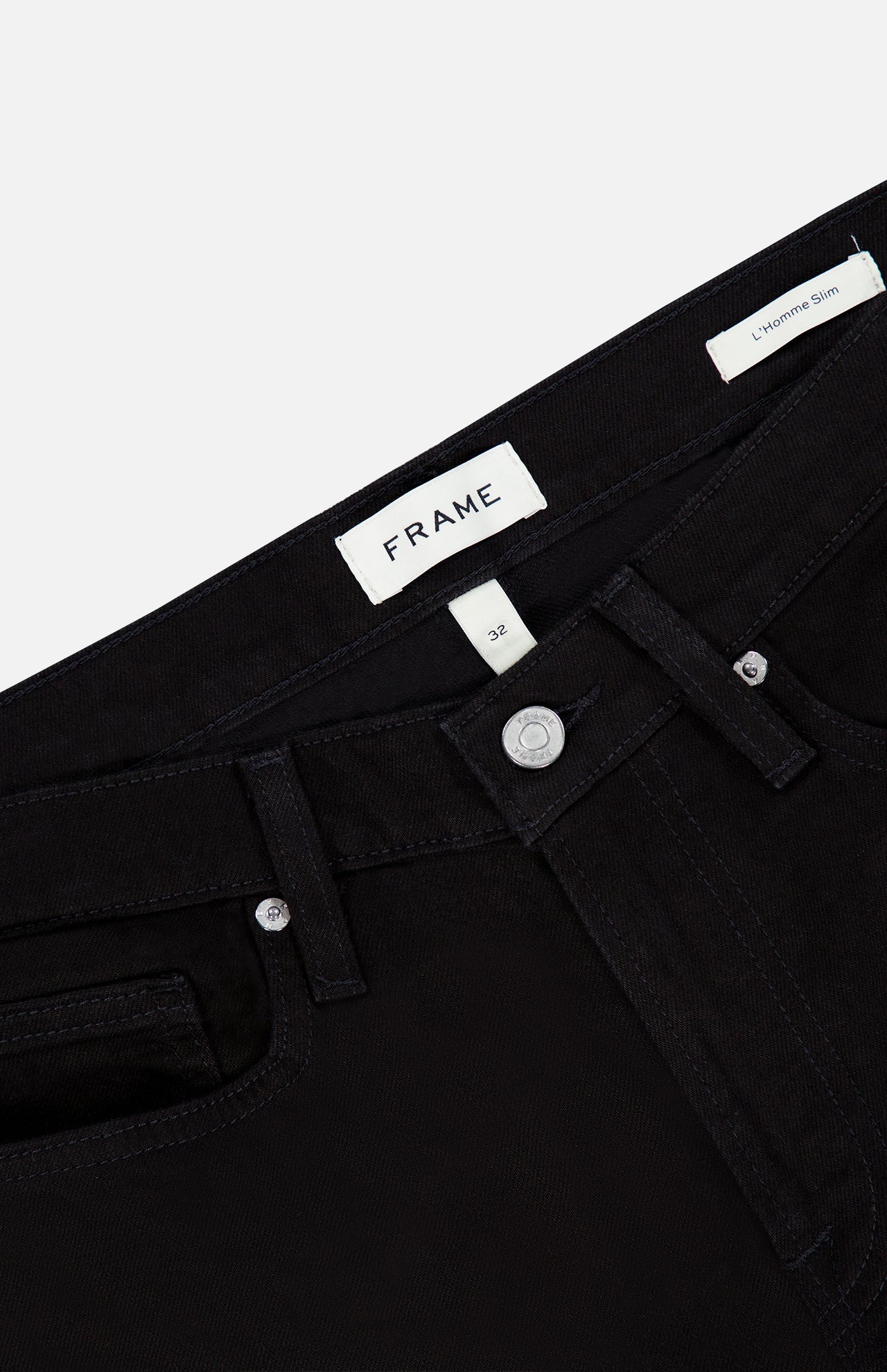 Close-up of the FRAME L’Homme Slim Pant in Noir, featuring a black denim design with a branded FRAME label on the waistband, silver button closure, front pocket, and a small tag reading “L’Homme Slim.” Crafted from premium stretch denim, these slim fit jeans combine modern style with exceptional comfort.