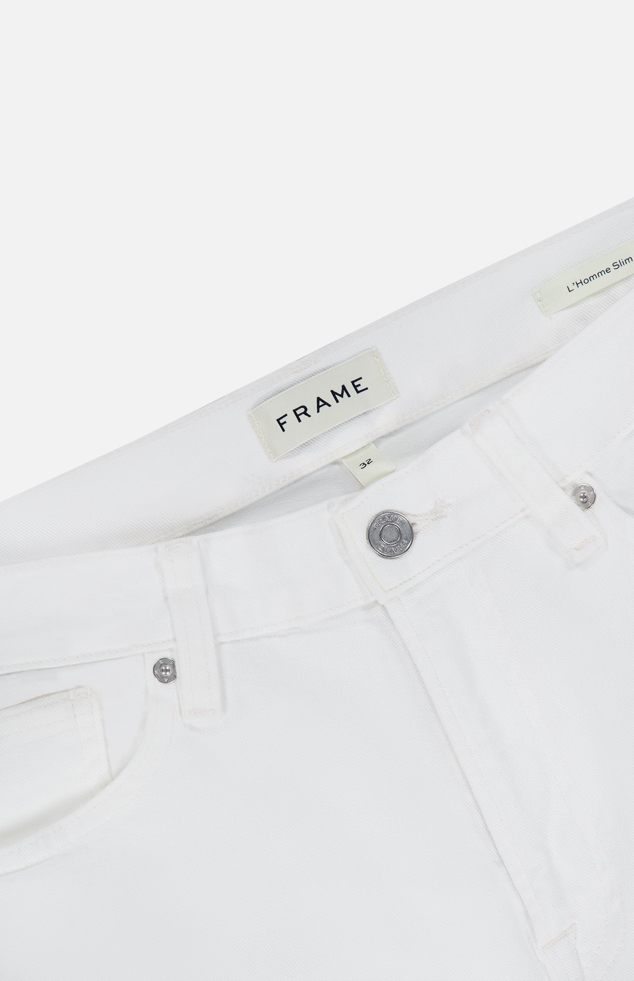 Close-up of the waistband and front pockets on the L’Homme Slim Pant in Whisper White by Frame, expertly crafted from premium stretch denim. Features include a metal button, an inner FRAME brand label, and a visible L’Homme Slim style tag.