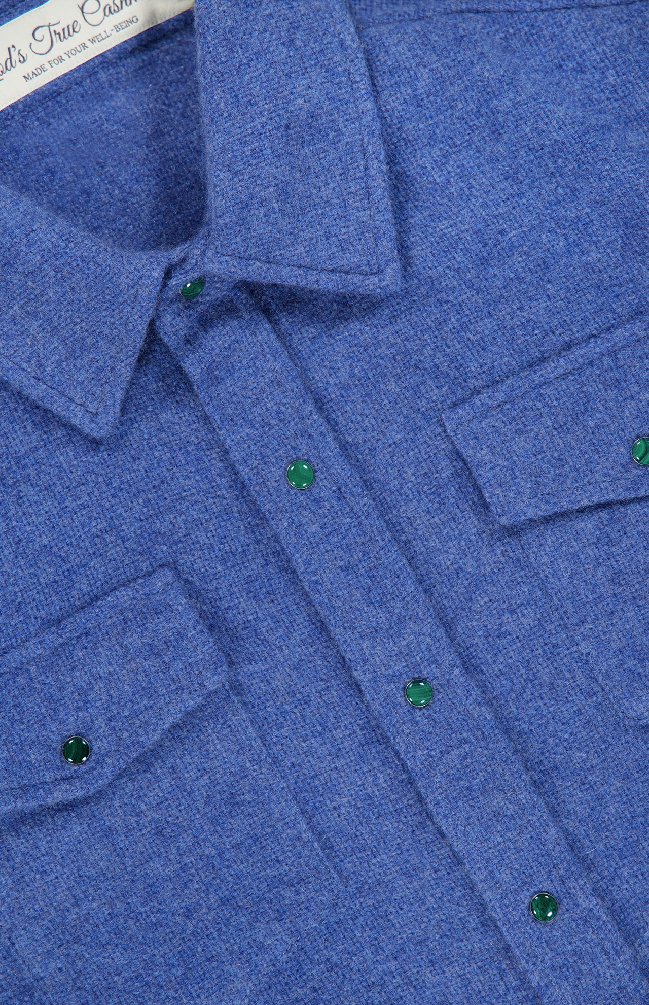 Close-up of the God’s True Cashmere Solid Cashmere Shirt featuring malachite-inspired green snap buttons, two chest pockets, and textured blue fabric. Part of a white label is visible at the top.