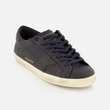 The GOLDEN GOOSE Super Star Leather Upper Sneaker is a dark navy low-top featuring a distinctive star design on the side, gold “GOLDENSTAR” text, black laces, and an off-white distressed sole. Styled against a plain white background, this contemporary pair reimagines classic navy sneakers with signature Golden Goose flair.