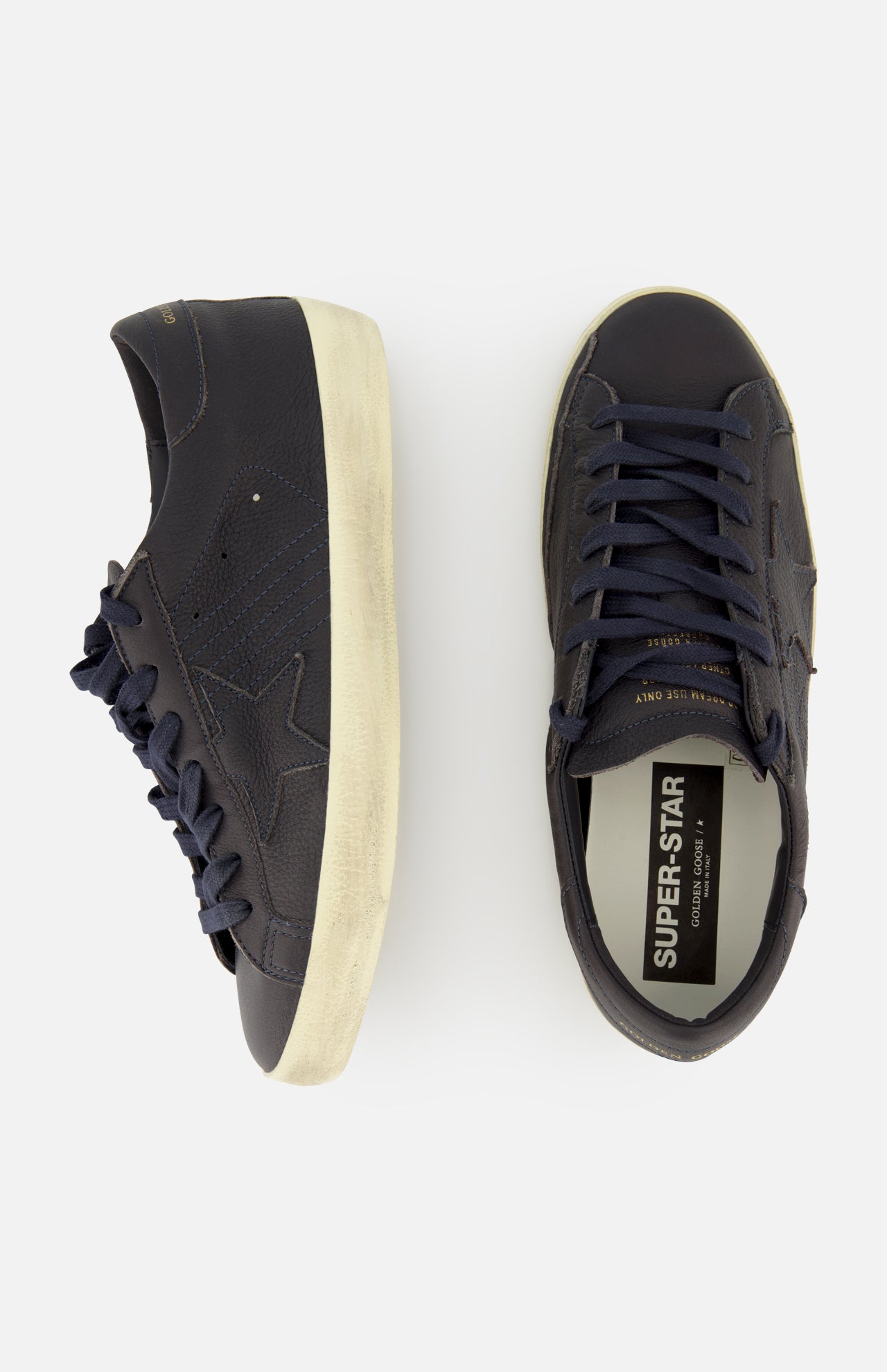 A pair of GOLDEN GOOSE Super Star Leather Upper Sneakers in navy blue with off-white, distressed soles is displayed from the side and top view, showcasing star accents and SUPER-STAR branding on the insole.