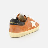 The GOLDEN GOOSE Super Star Suede Upper Toe And Tongue Sneaker features a brown suede upper, a distinctive white star accent on the side, beige laces, and a black heel tab with bold yellow text. Its signature scuffed sole completes the look, all showcased against a plain white background.