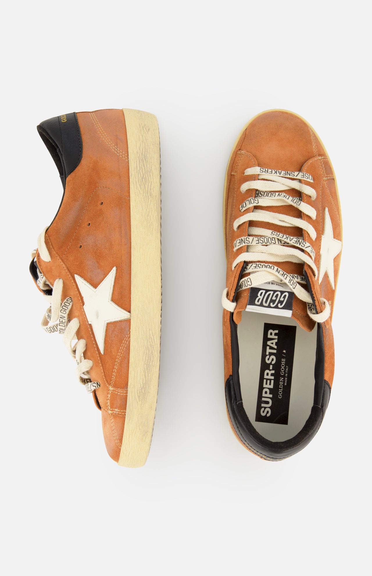 A pair of GOLDEN GOOSE Super Star Suede Upper Toe And Tongue Sneakers in tan, featuring white stars, black heel tabs, and cream-colored laces. The sneakers are finished with the brand's signature scuffed sole for a distinctive look. One shoe is shown from the side while the other is viewed from above, revealing "SUPER-STAR" on the insole.