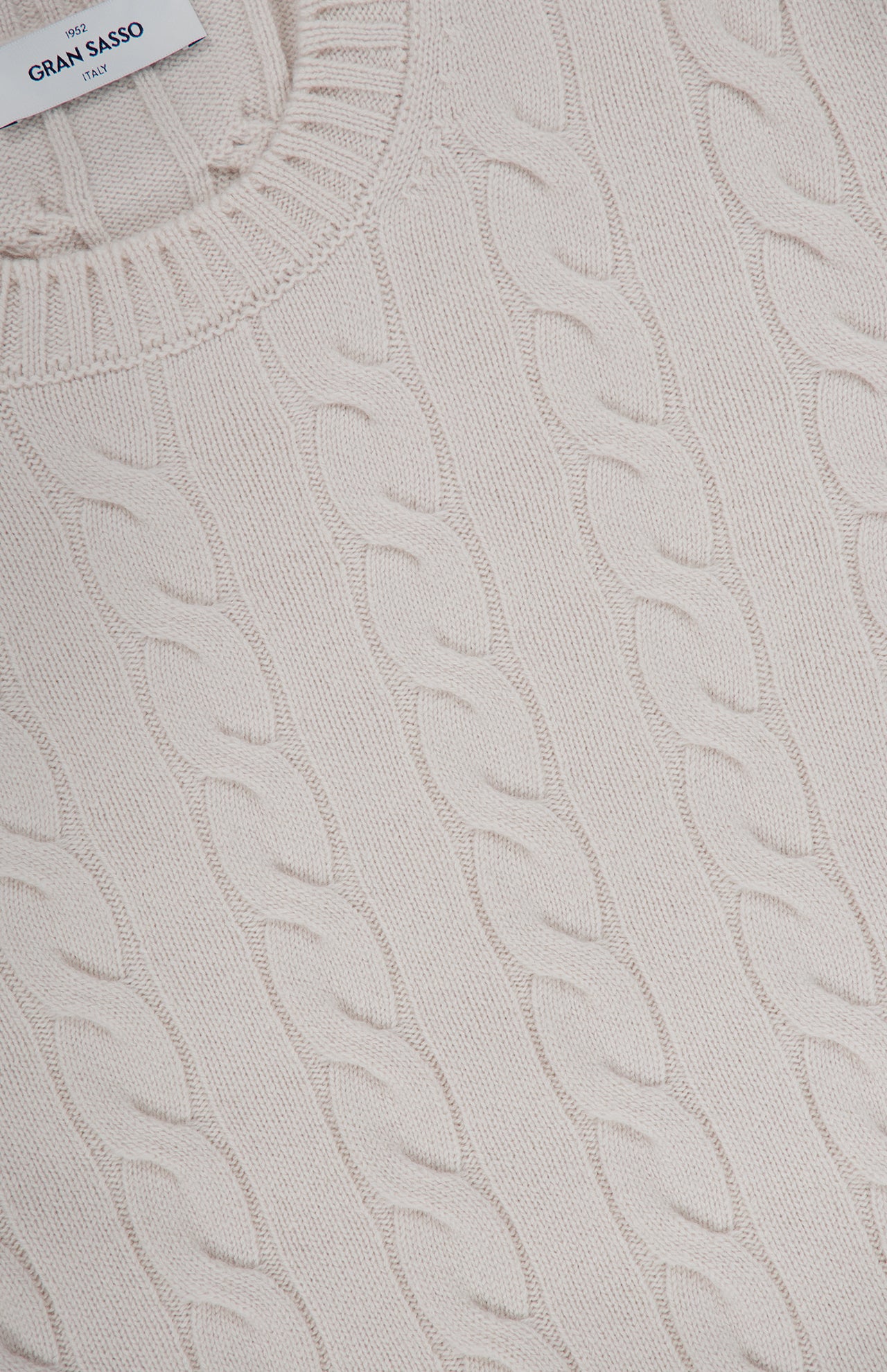 Close-up of the Cable Knit Crewneck Sweater by GRAN SASSO. This beige pullover features a classic round neckline and displays the GRAN SASSO ITALY label at the collar. The timeless braided cable knit pattern showcases its refined wool-blend construction.