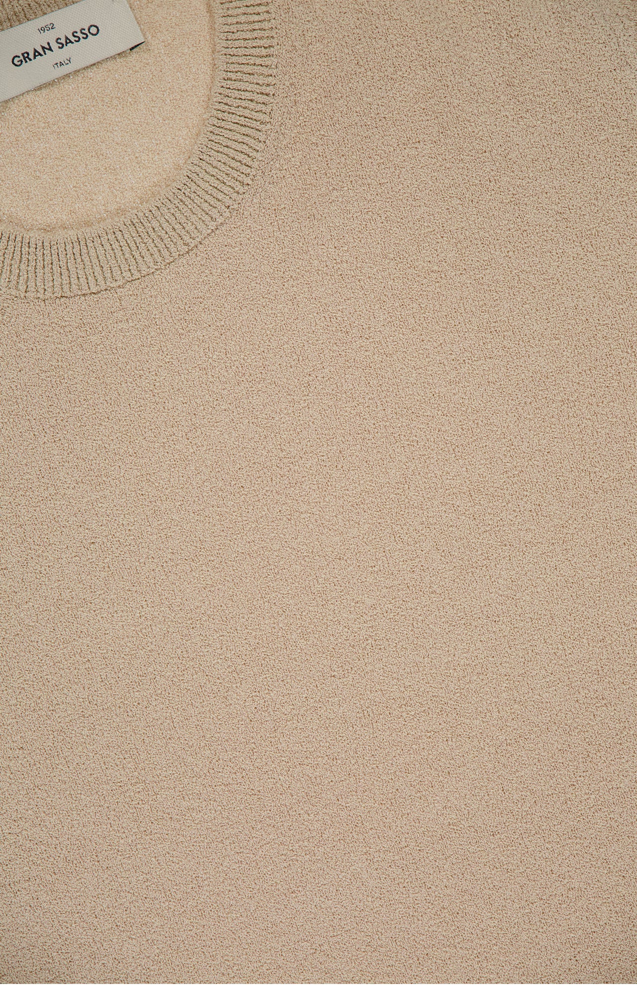 Close-up of a beige French Terry Short Sleeve Crewneck Sweater by GRAN SASSO, featuring a ribbed neckline and a white label in the top left corner with the brand name.