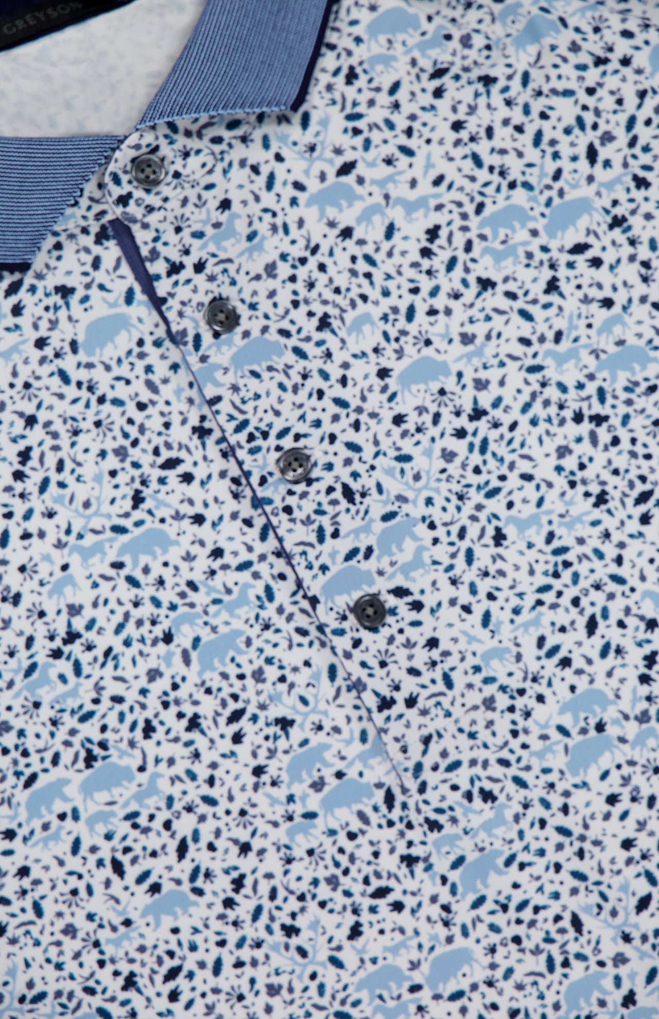 Close-up of the GREYSON Buffalo Solider Polo Shirt, featuring blue and white fabric with small animal silhouettes and abstract shapes. It offers UV protection and has a blue collar with four buttons.