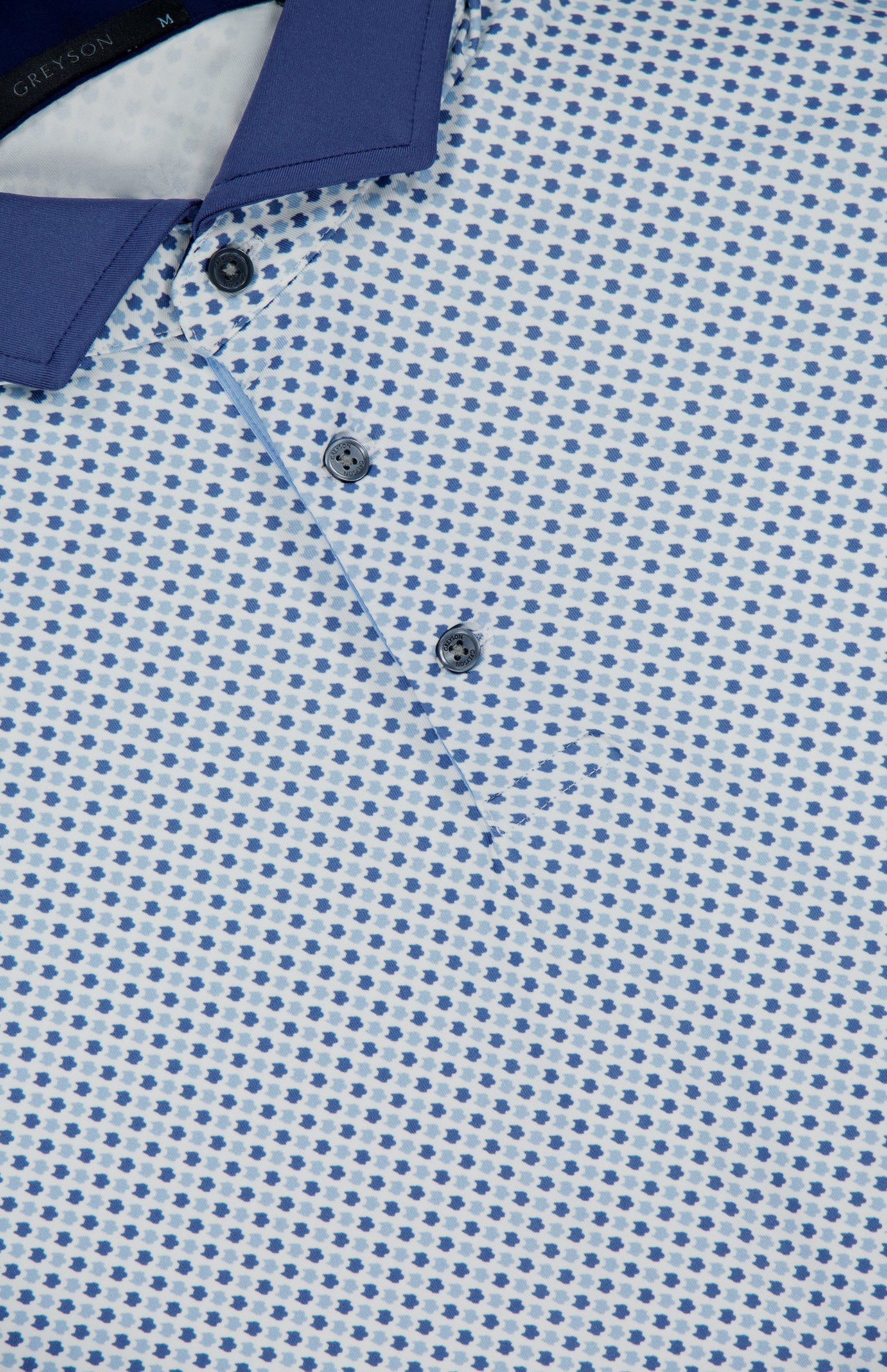 Close-up of the GREYSON Ironwood Polo Shirt, showcasing a blue and white geometric dot pattern, three dark buttons, a solid blue collar, and moisture-wicking fabric for comfort.