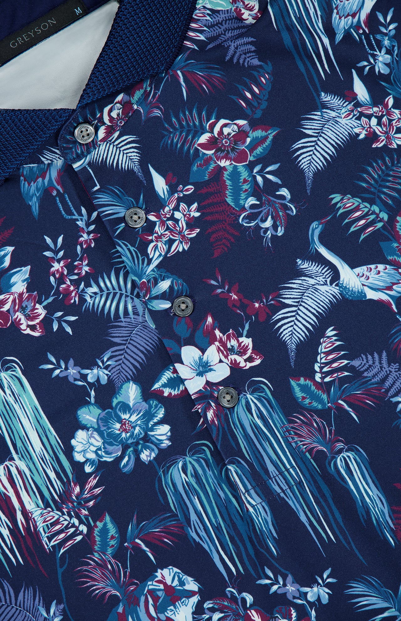 A close-up of the GREYSON Night Lurk Polo Shirt in navy blue, showcasing a vibrant tropical print, moisture-wicking fabric, and button-down collar. The label inside the collar highlights its blend of bold style and performance innovation.