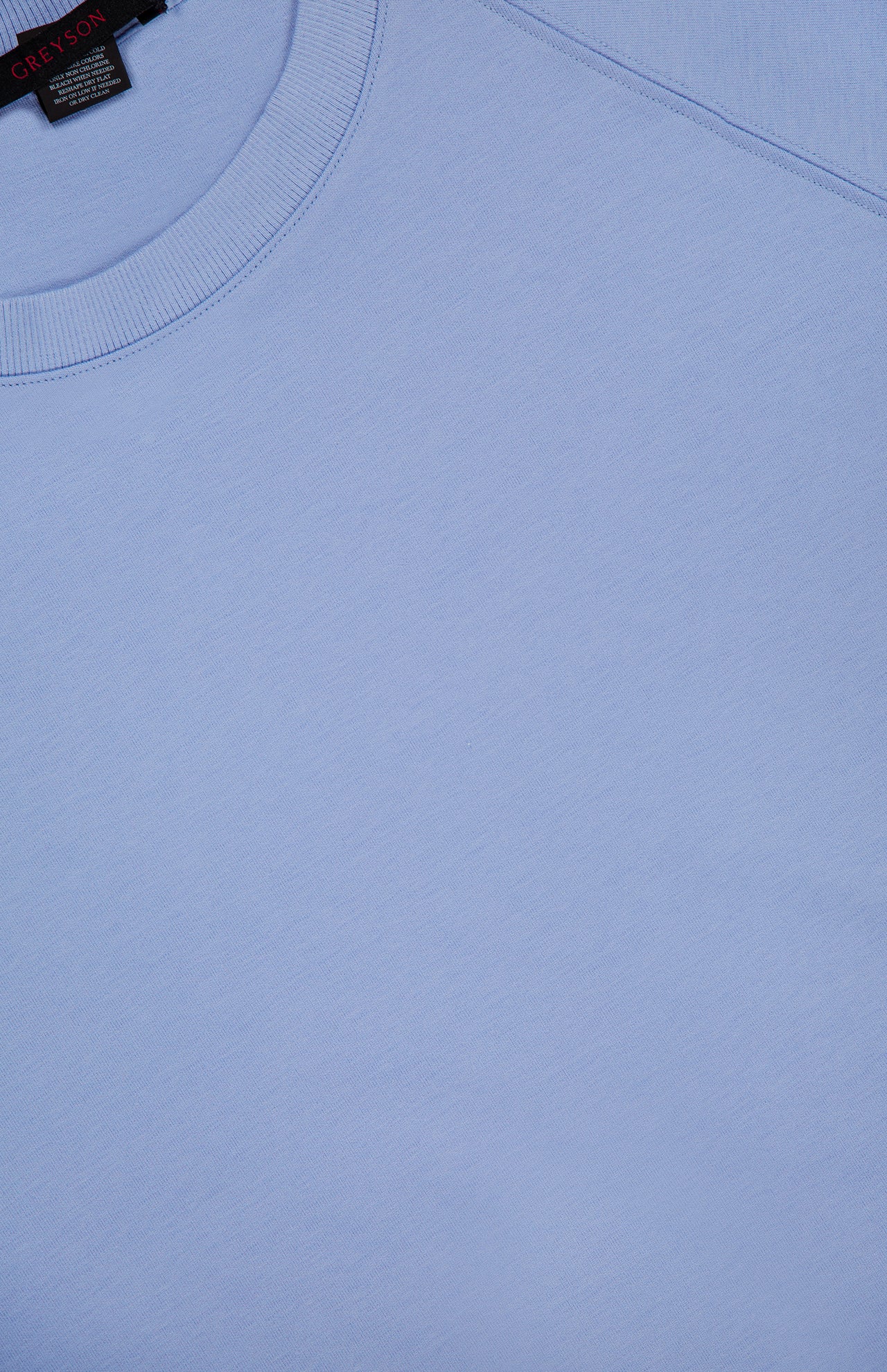 A close-up of the Ojai Cropped Crewneck Sweatshirt by GREYSON in light blue highlights the soft cotton-blend fabric, crew neck collar, shoulder seam, and a small section of its black tag with red text.