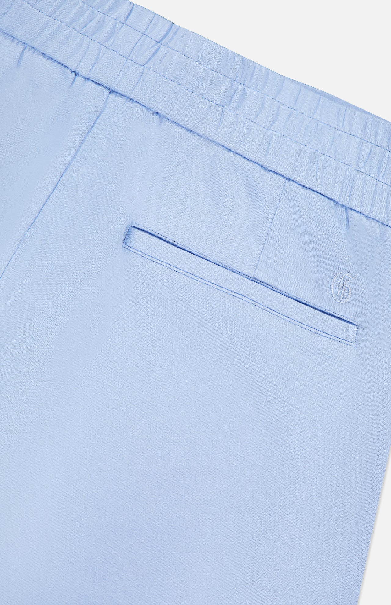 Close-up of the Ojai Trouser Short by GREYSON in light blue, showing an elastic waistband, welt pocket, and a small embroidered G logo above the right-side pocket.
