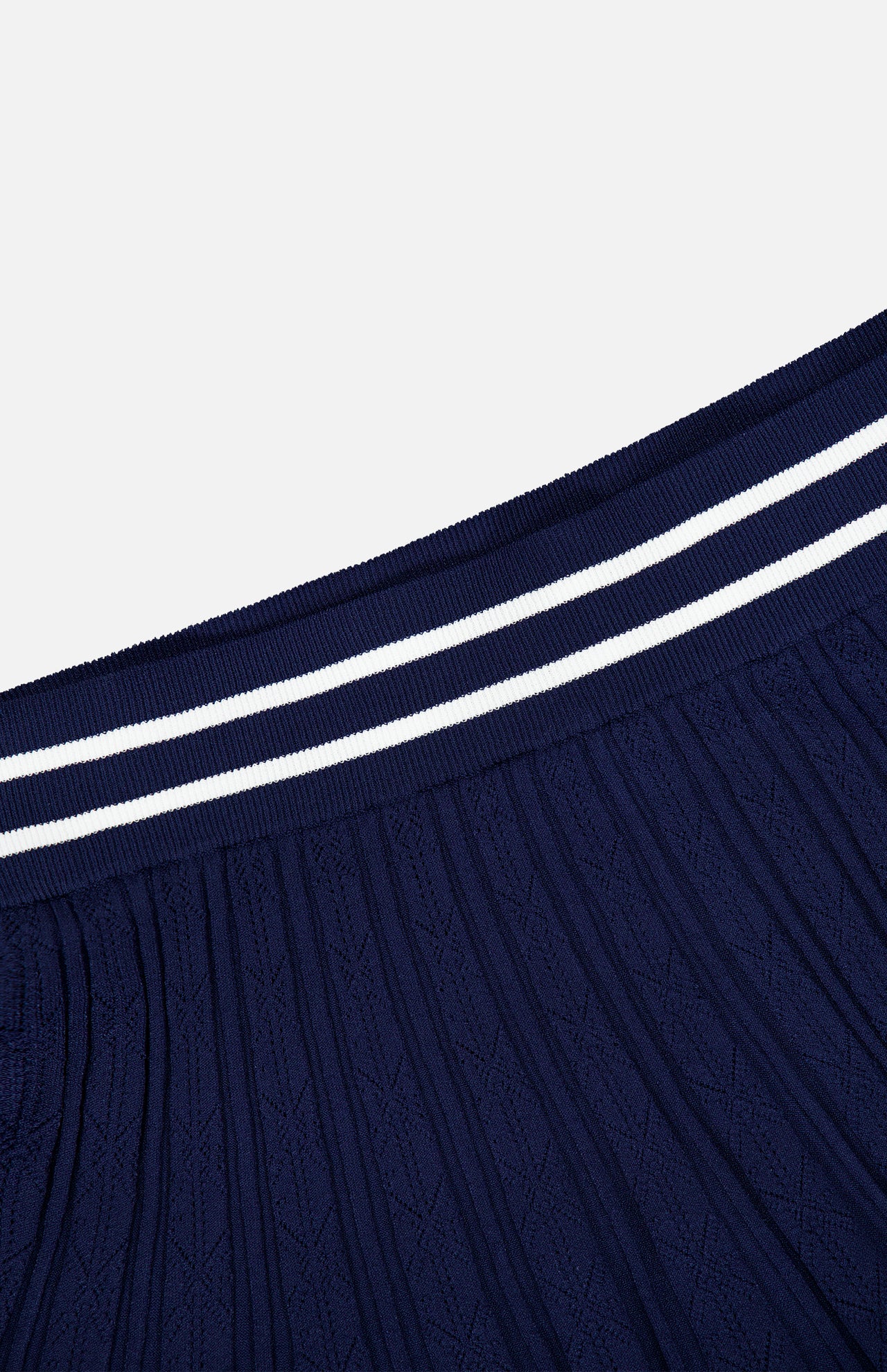 The GREYSON Pointelle Mini Skirt is shown in close-up against a plain white background. Crafted from pleated dark blue fabric, it features a ribbed waistband with two parallel white stripes. The quick-dry material and built-in undershort deliver comfort and practicality for all-day wear.