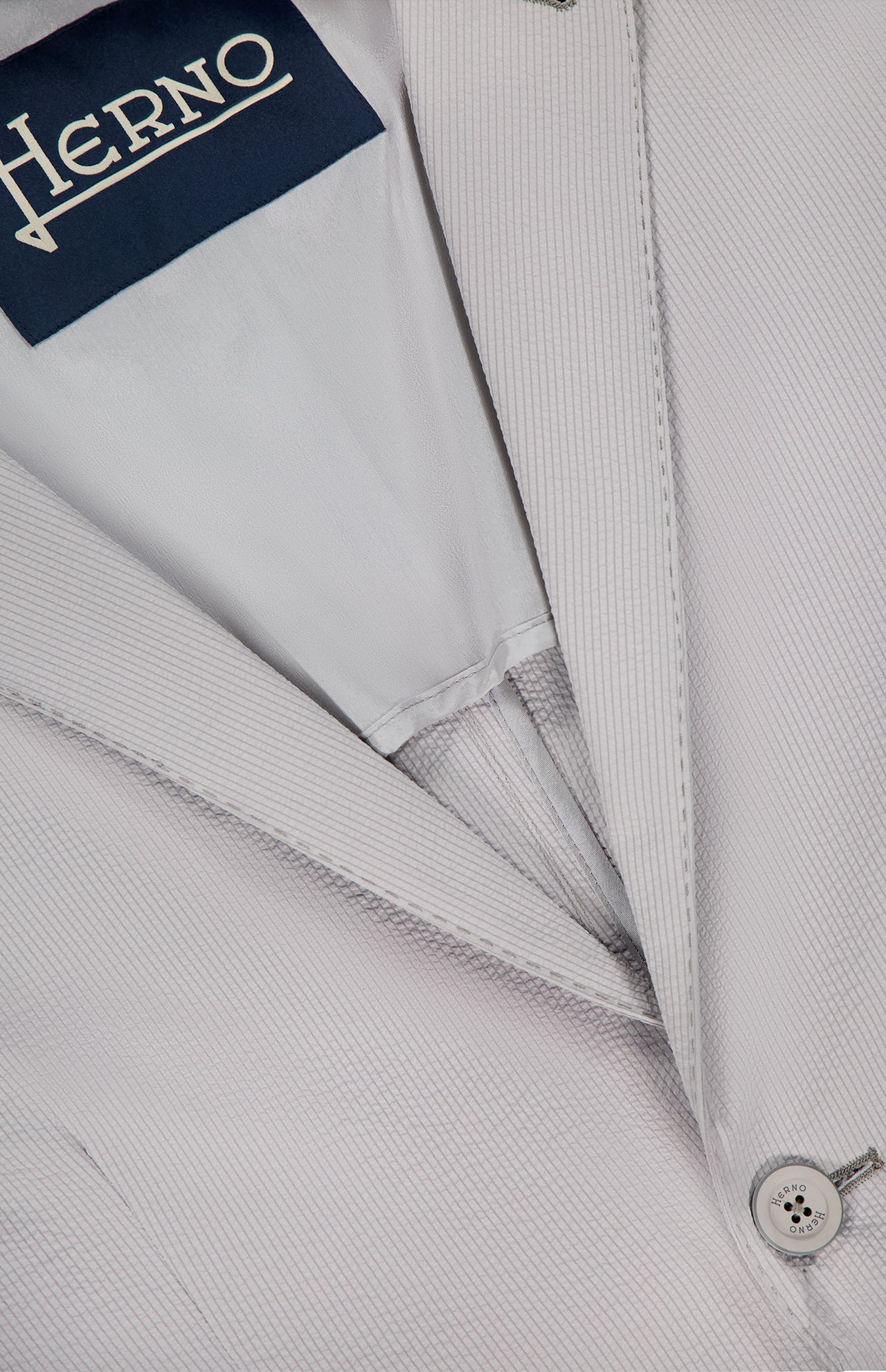 Close-up of the HERNO Seersucker 2 Button Blazer for men, shown in light grey textured fabric with visible button, lapel, and navy blue interior HERNO label.