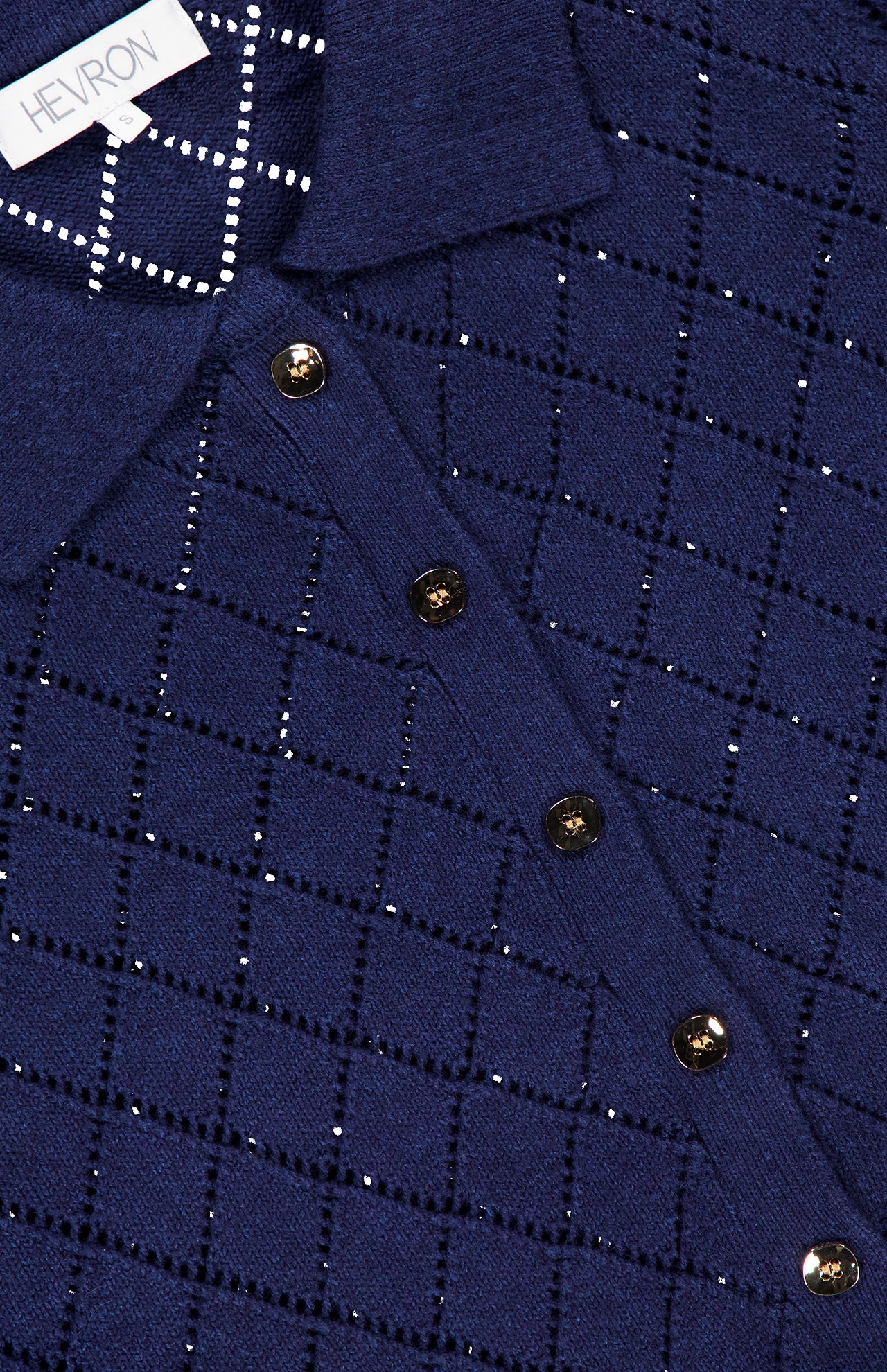 A close-up of the Arlet Diamond Pointelle Top by HEVRON shows its navy blue knit, collar, gold buttons, and diamond pointelle pattern. The white HEVRON label is visible near the neckline.