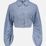 The Raquel Pleated Sleeves Top by HEVRON features blue stripes, a cropped button-up design, collar, chest pocket, and exaggerated pleated sleeves for a modern touch.