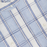 Close-up of the Eva Linen Cropped Top by HEVRON, in light blue and white plaid with dark blue lines. Features white buttons, soft textured linen fabric, and a partially visible shirt label in the top left corner.