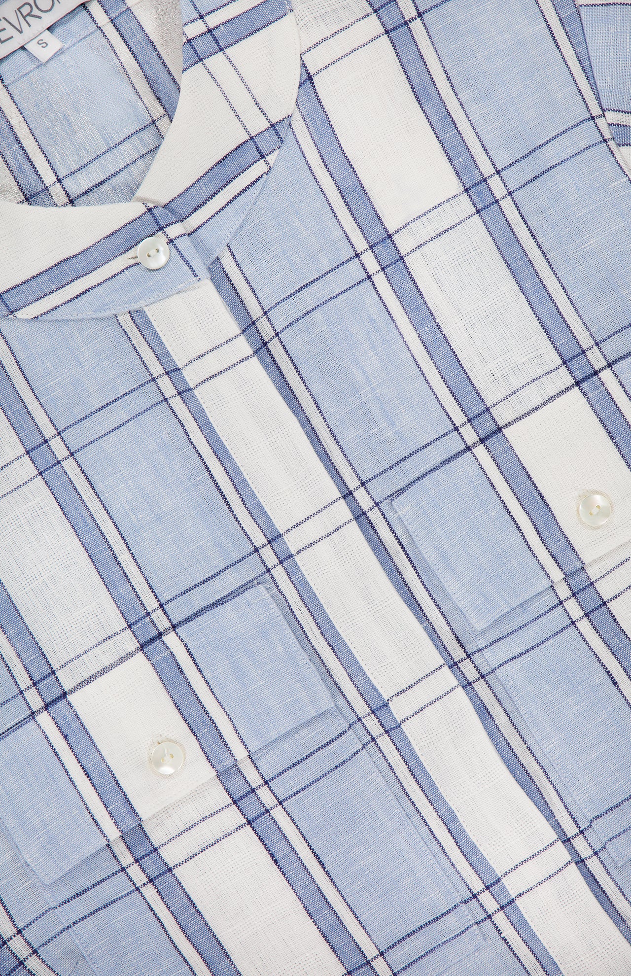Close-up of the Eva Linen Cropped Top by HEVRON, in light blue and white plaid with dark blue lines. Features white buttons, soft textured linen fabric, and a partially visible shirt label in the top left corner.