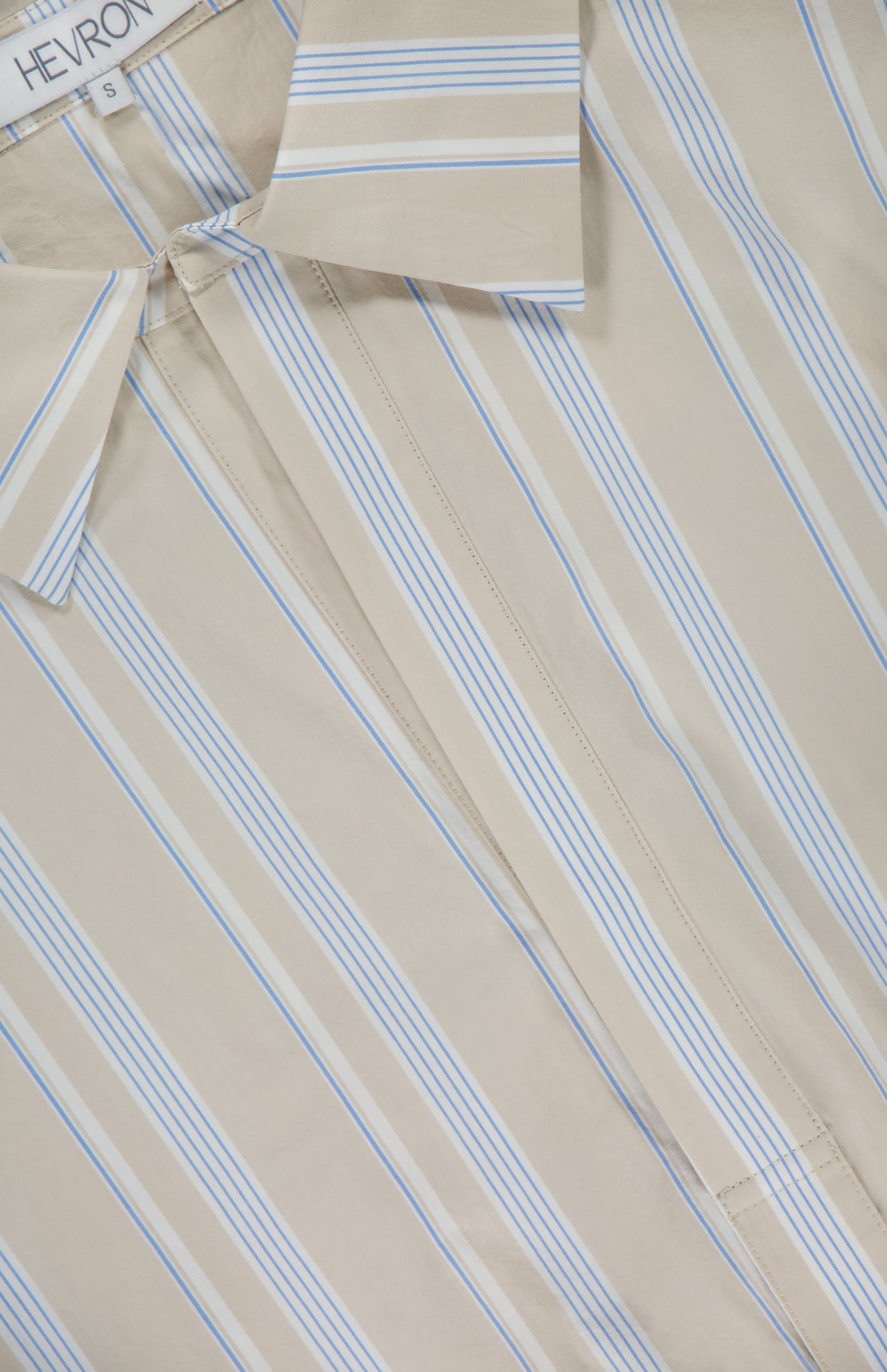 A close-up of the Paola Front Tuck Top by HEVRON shows beige fabric with blue and white vertical stripes, a visible size S tag, the collar, part of the button placket, and a hint of back button detail.