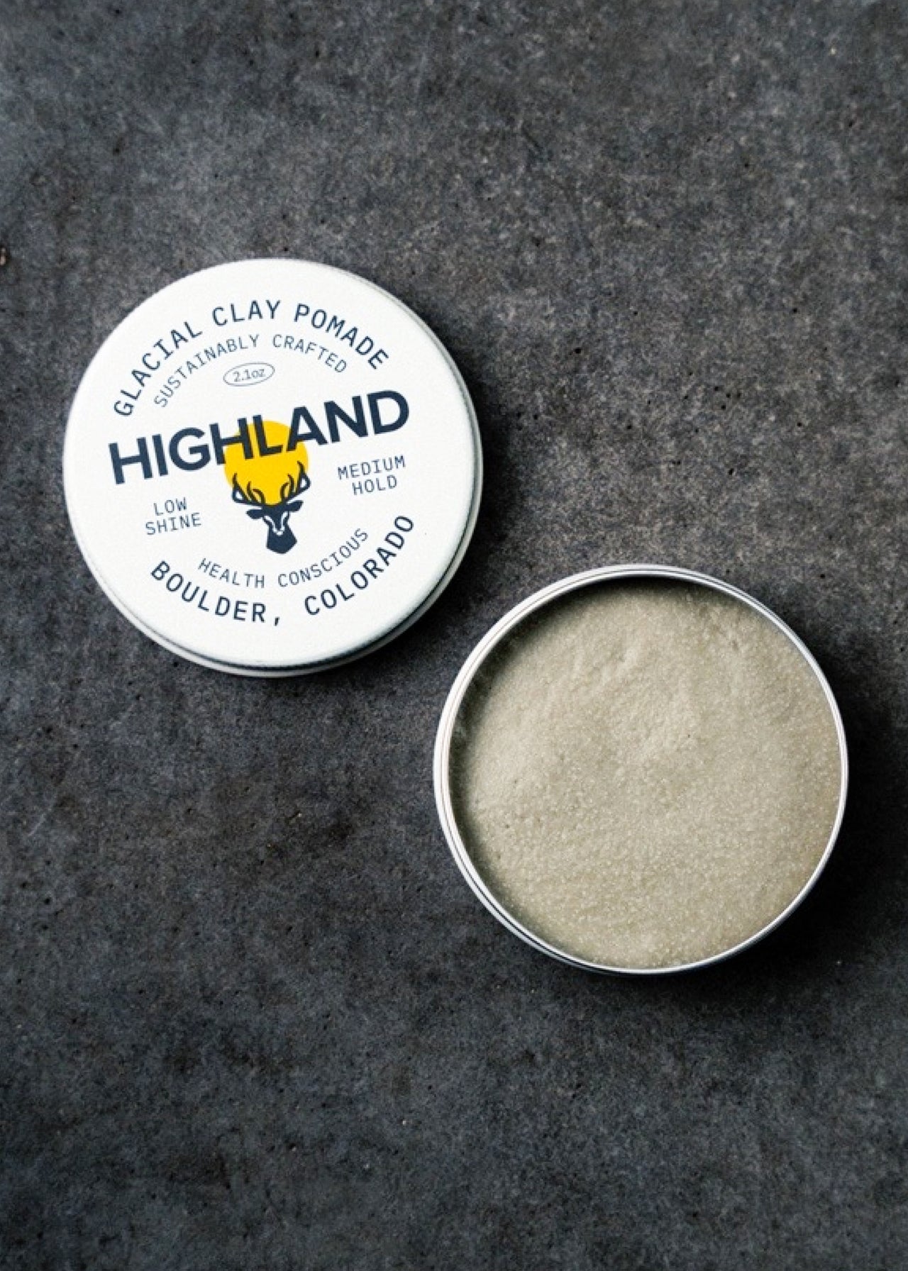 A round tin of HIGHLAND STYLE CO. Glacial Clay Pomade with the lid off reveals light beige clay inside. The gray textured lid displays brand and product details—crafted with natural ingredients for healthy scalp care.