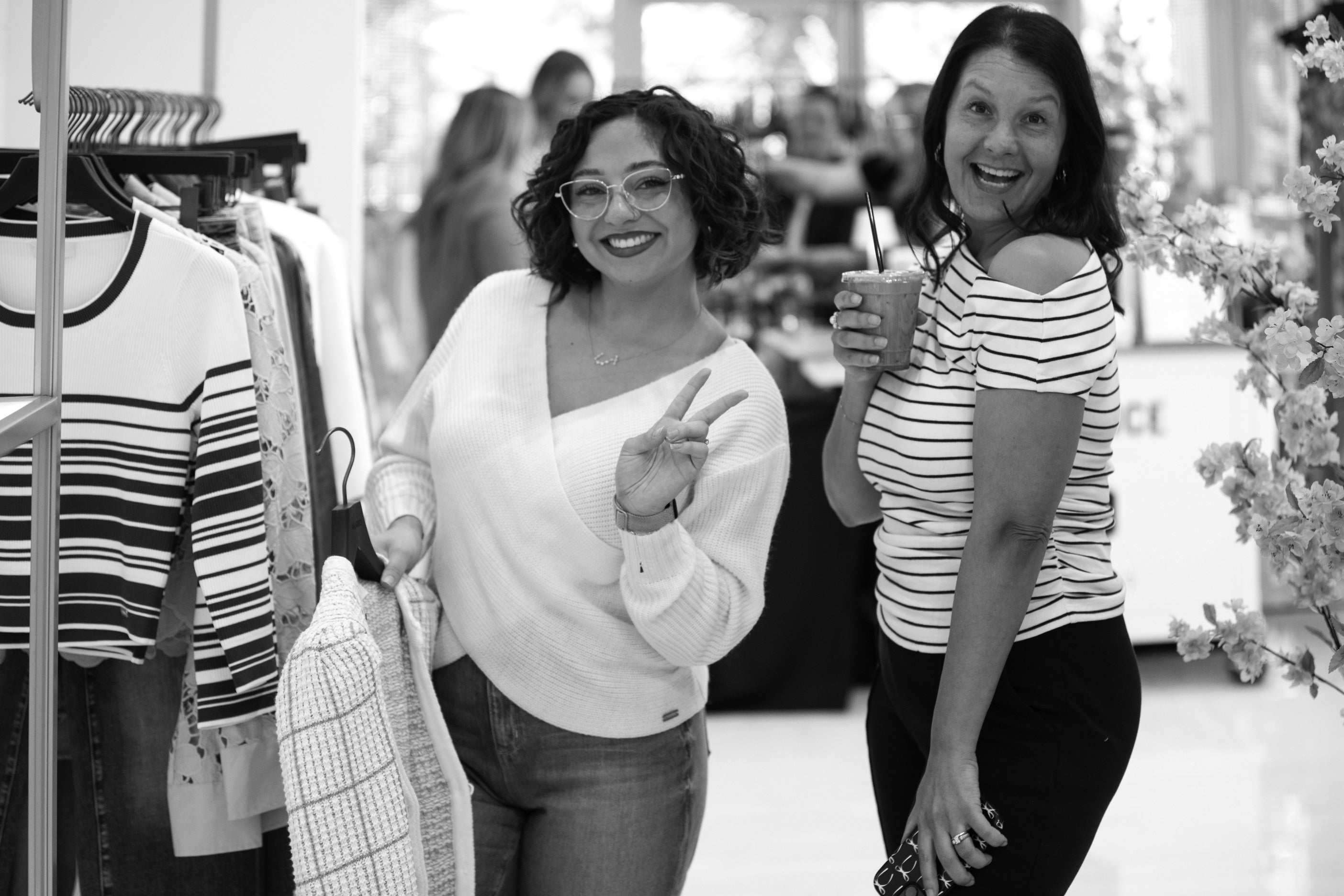 Two women posing in a clothing store with a rack of clothes in the background.