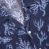 A close-up of the ISAIA Corallina Print Camp Shirt shows its navy linen fabric with a light blue coral pattern, white buttons, and a red collar label, highlighting the shirt's soft, textured look.