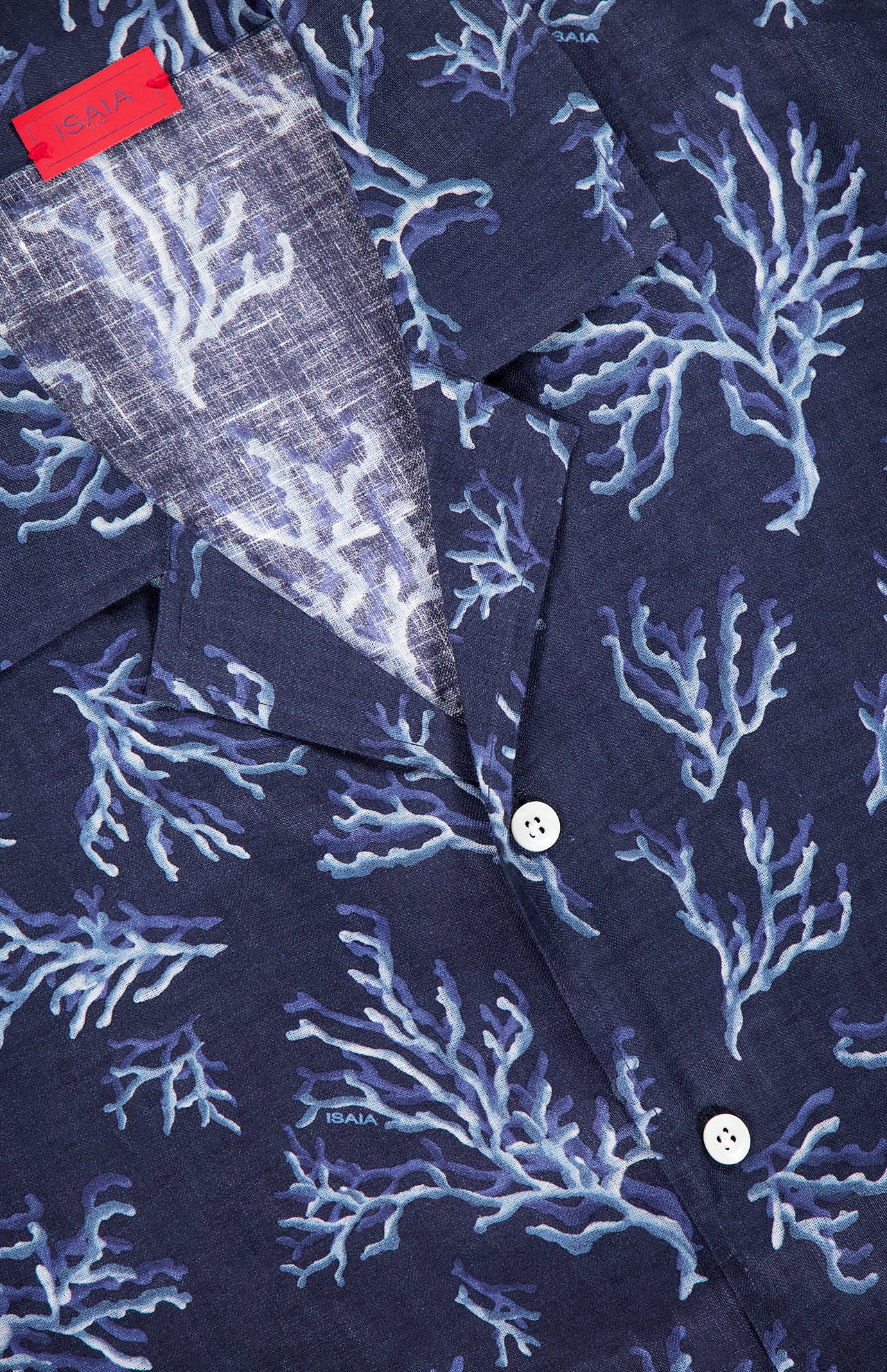 A close-up of the ISAIA Corallina Print Camp Shirt shows its navy linen fabric with a light blue coral pattern, white buttons, and a red collar label, highlighting the shirt's soft, textured look.