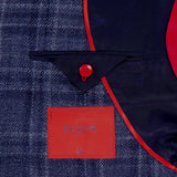 Close-up of the ISAIA Galles Over Check Rustic Sportcoat interior, featuring a red-edged pocket flap, red button, and red label on navy lining. The blue plaid fabric in the background highlights its made-in-Italy craftsmanship.