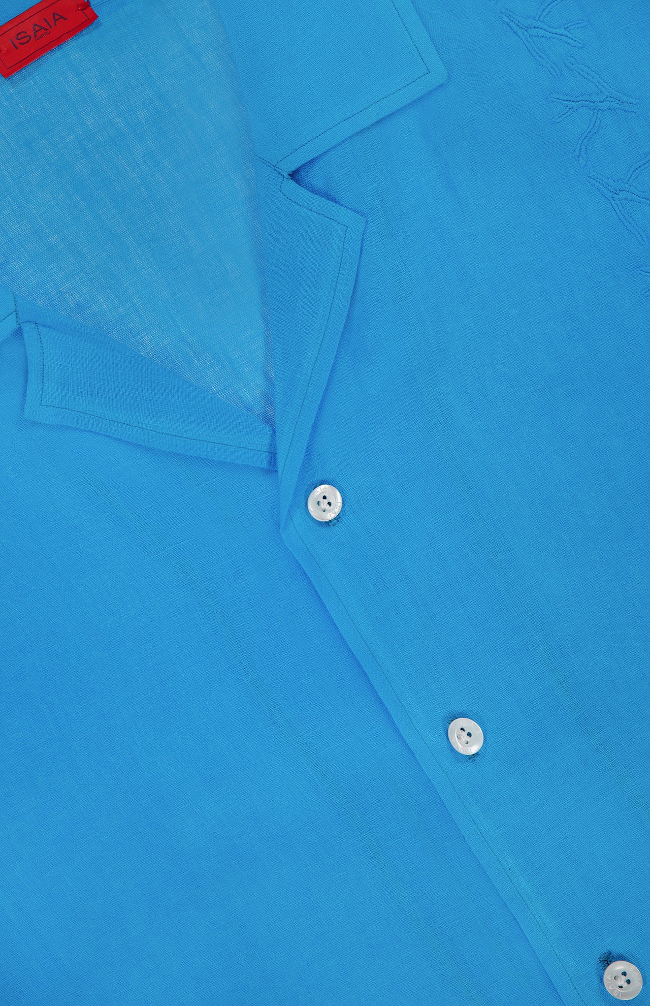 Close-up of the ISAIA Linen Coral Embroidery Shirt in bright blue, featuring a collar, white buttons, subtle coral embroidery, and a red brand tag inside the top left collar for a relaxed vacation look.