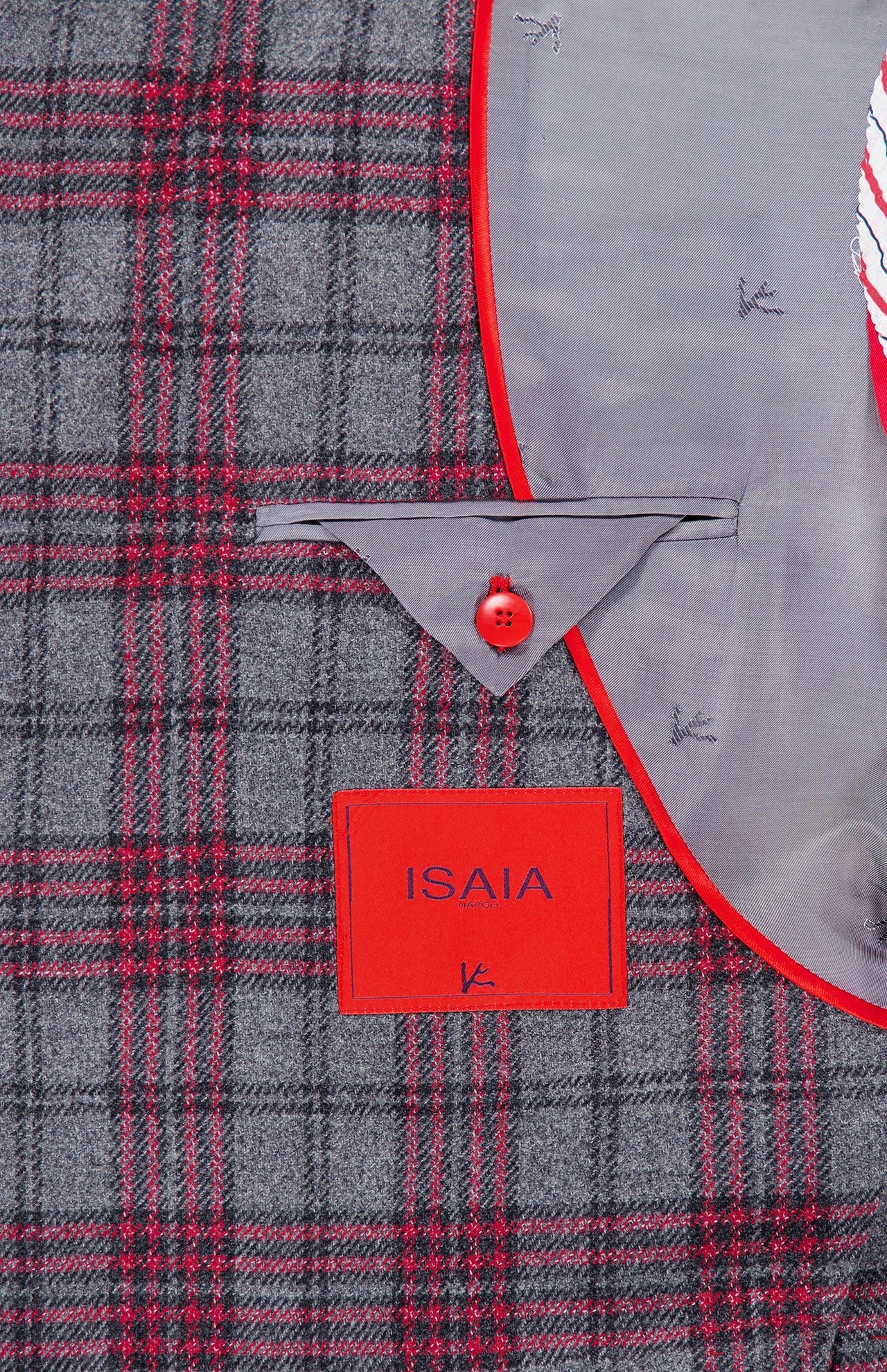 Close-up of the Over Checkered Cashwool Sportcoat by ISAIA, an Italian-made jacket in a grey and maroon plaid pattern. It features a red-bordered pocket, signature red button, and a distinctive red rectangular ISAIA label. The light gray lining is adorned with finely embroidered fish motifs, showcasing the meticulous craftsmanship of this wool-cashmere sportcoat.
