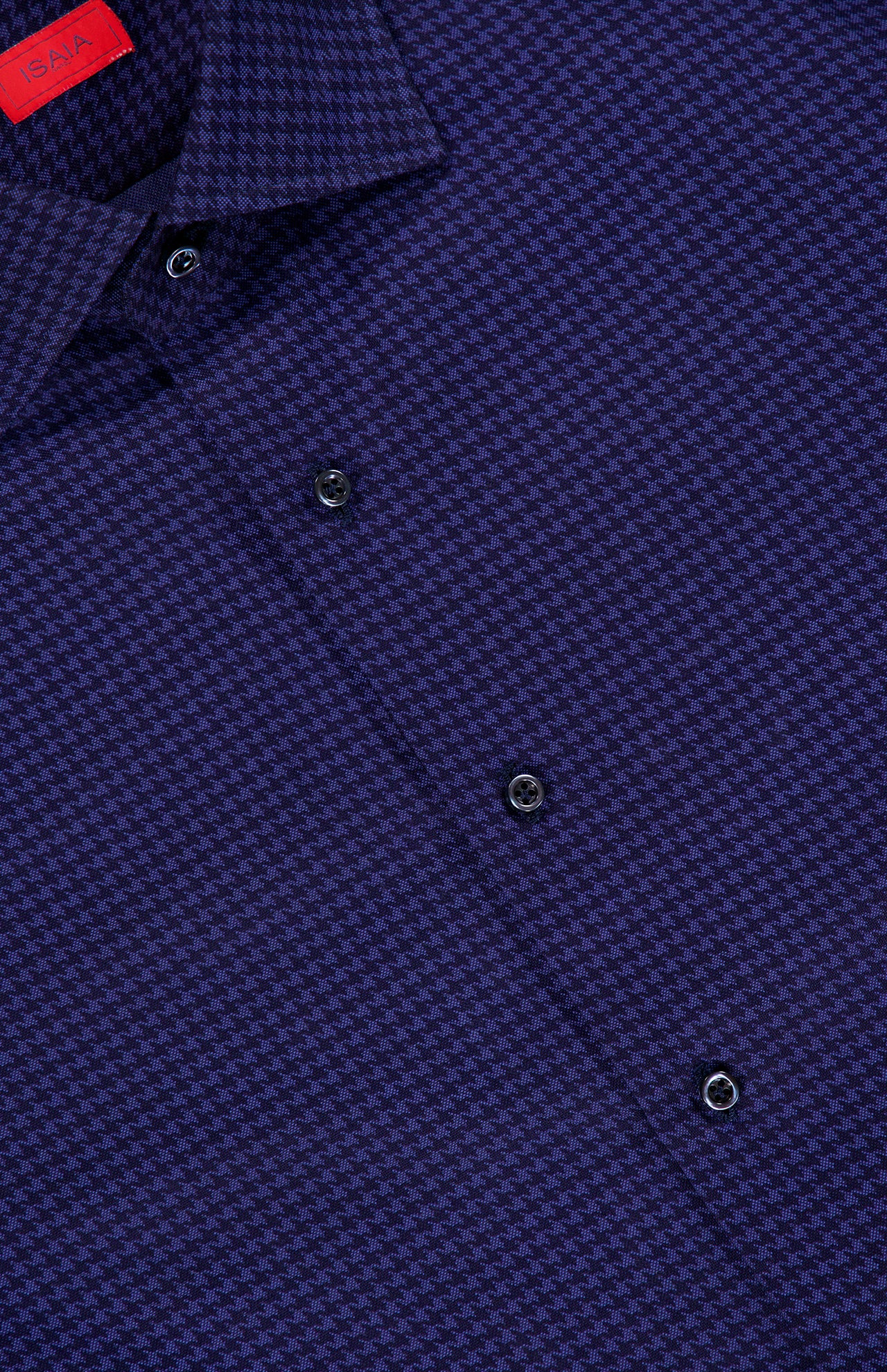Close-up of the ISAIA Print Oxford Dress Shirt, featuring a blue and black micro houndstooth pattern with black buttons. The collar and a red ISAIA label are partially visible in the upper left corner, showcasing refined detailing characteristic of Italian dress shirts.